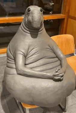 Grey seal-like statue sitting in a chair with its hands folded, the waiting meme