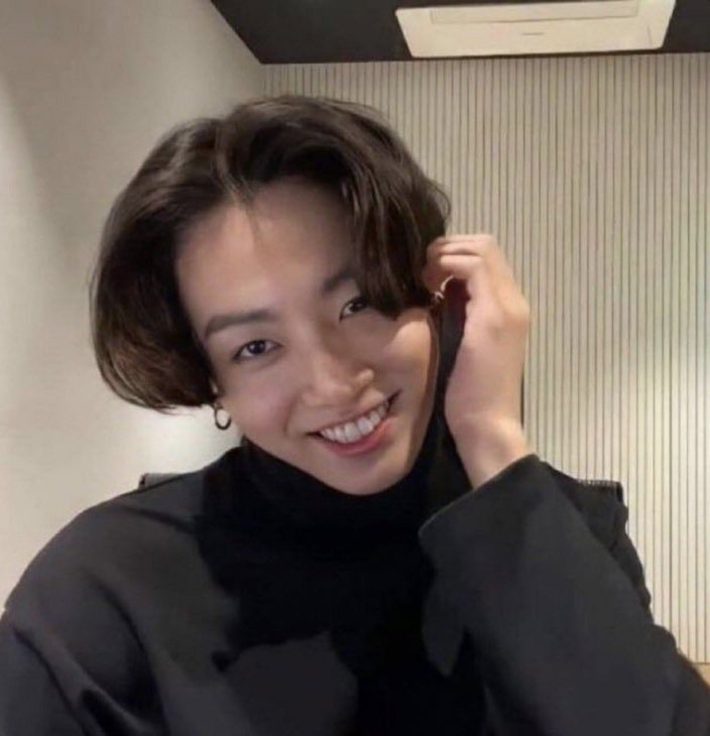 Person in a black turtleneck beaming while tucking their hair back