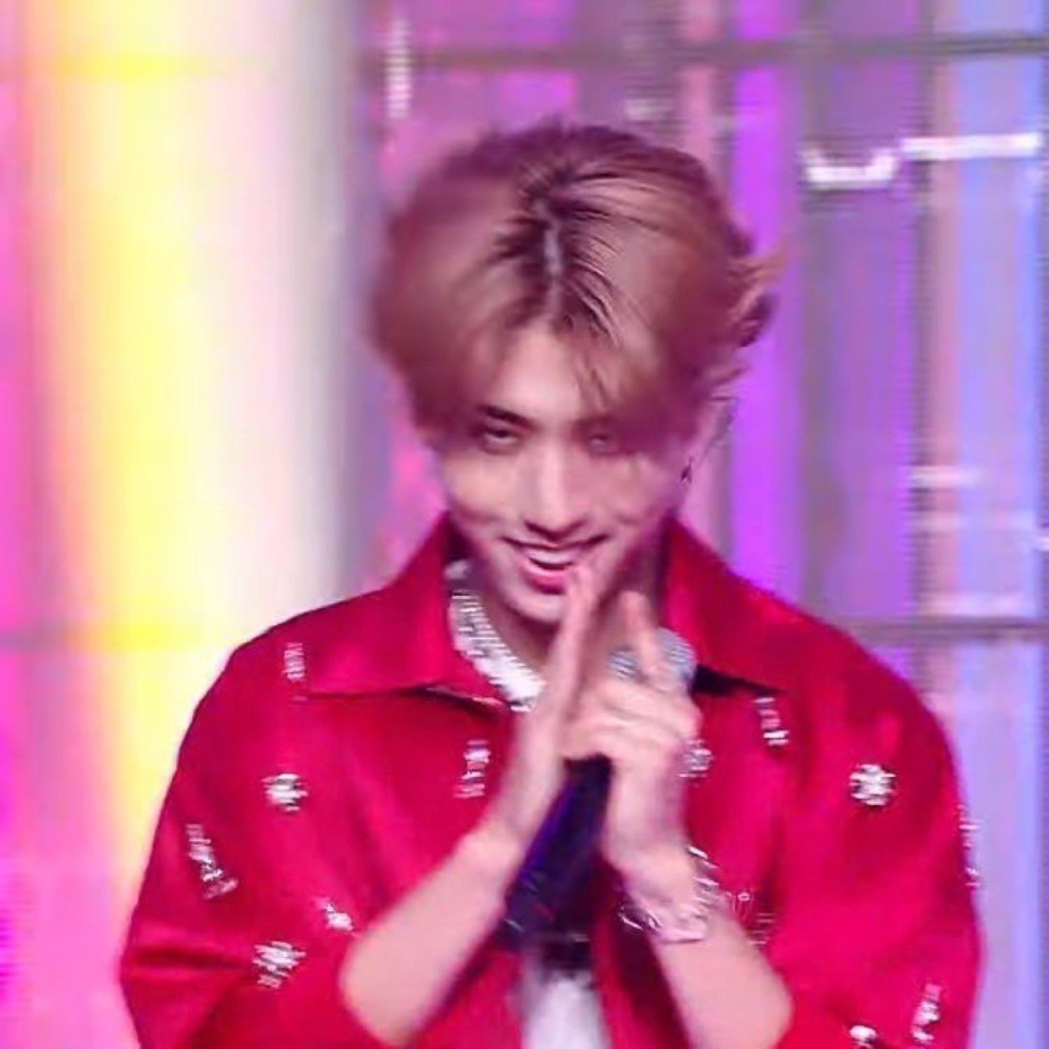K-pop idol in a red shirt on stage, hands clasped with a shy grinning look