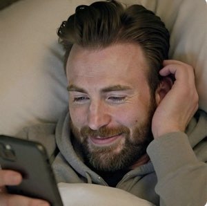 Bearded man lying in bed grinning softly at his phone