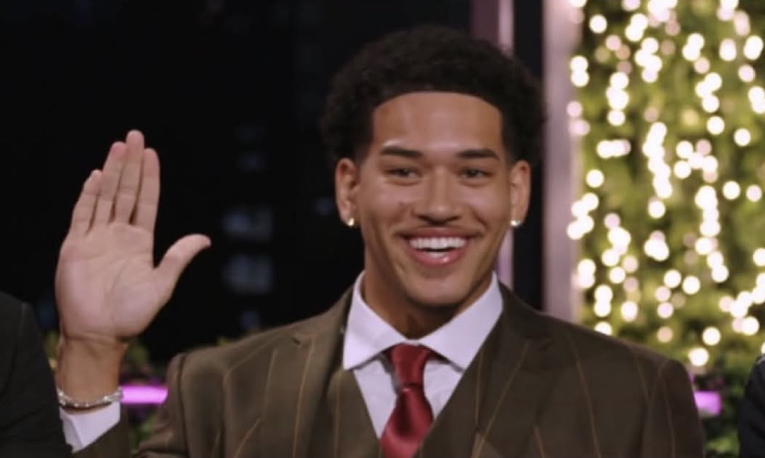 Love Island USA's Corbin in a suit raising his hand with a smile