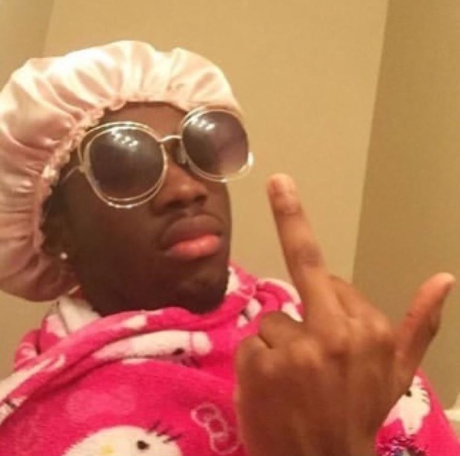 Man in a bonnet, huge sunglasses and Hello Kitty robe throwing up both middle fingers