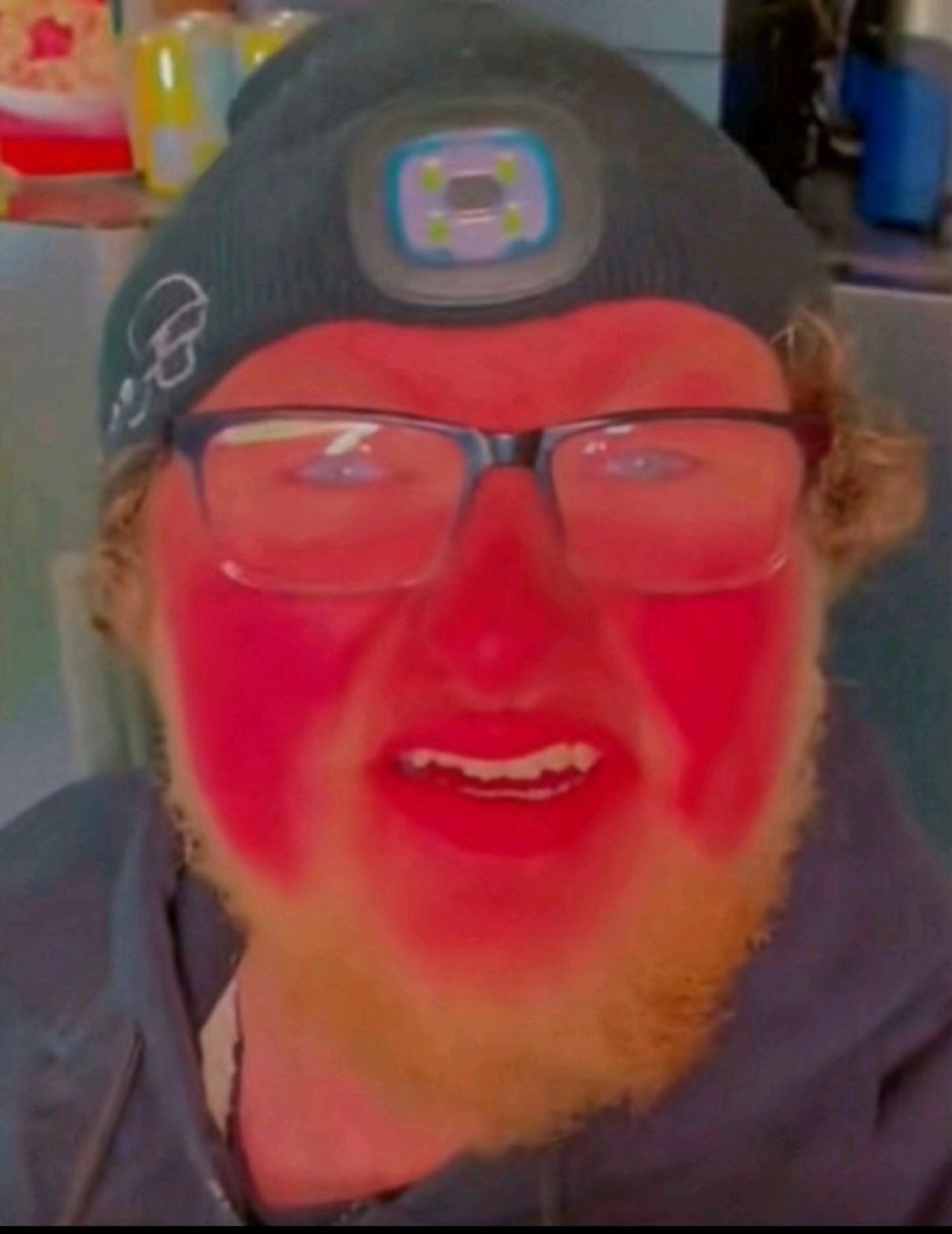Man with red-flushed face filter grinning menacingly with a headlamp hat