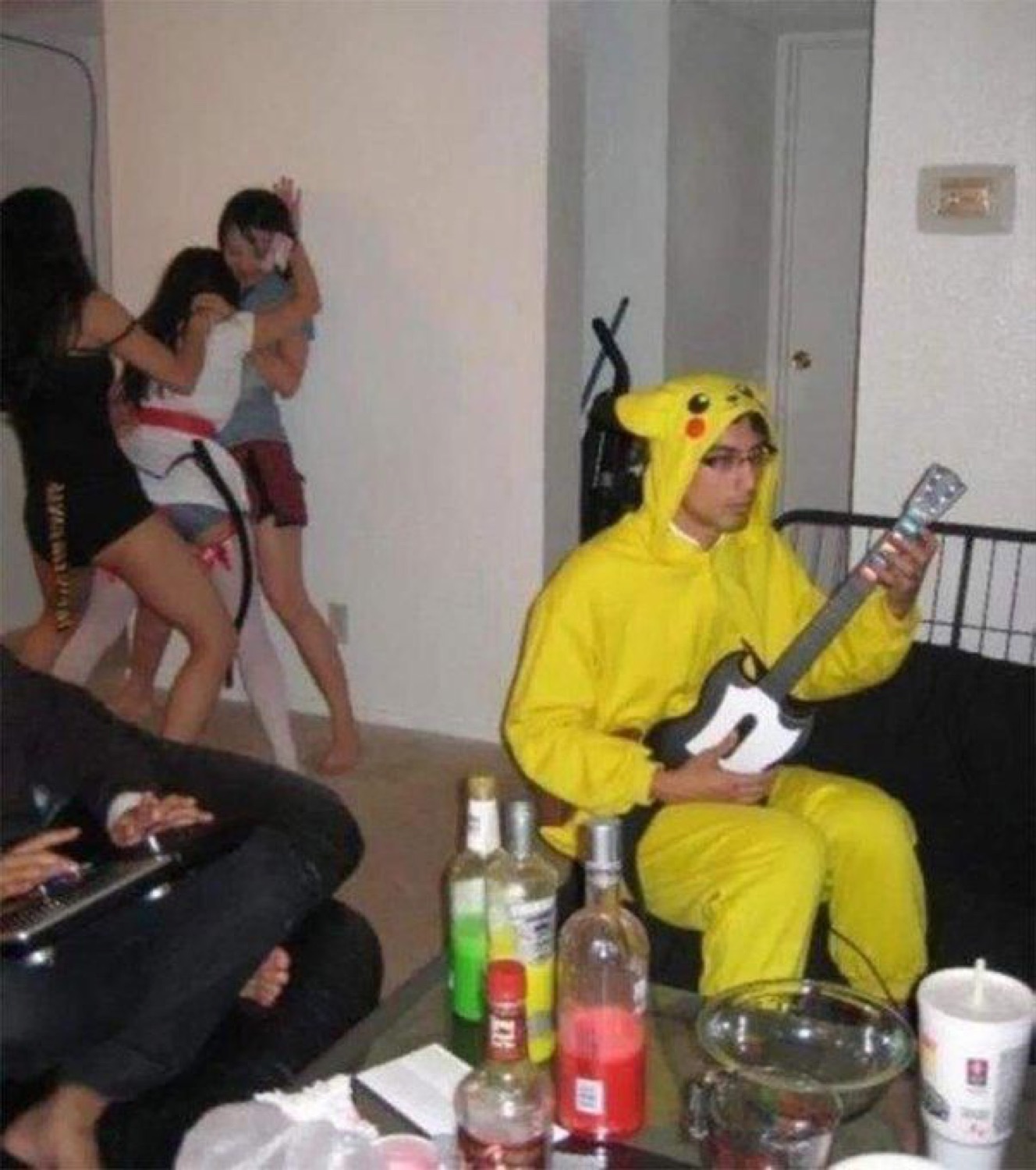 Guy in a full Pikachu onesie playing Guitar Hero alone at a party while others mingle