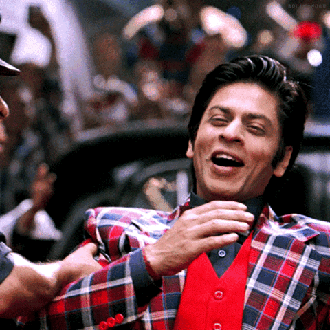 Shah Rukh Khan in a plaid jacket laughing with his hand on his chest