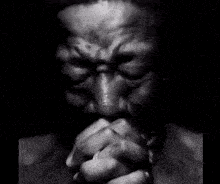 Man in black and white with his hands clasped in prayer, looking up