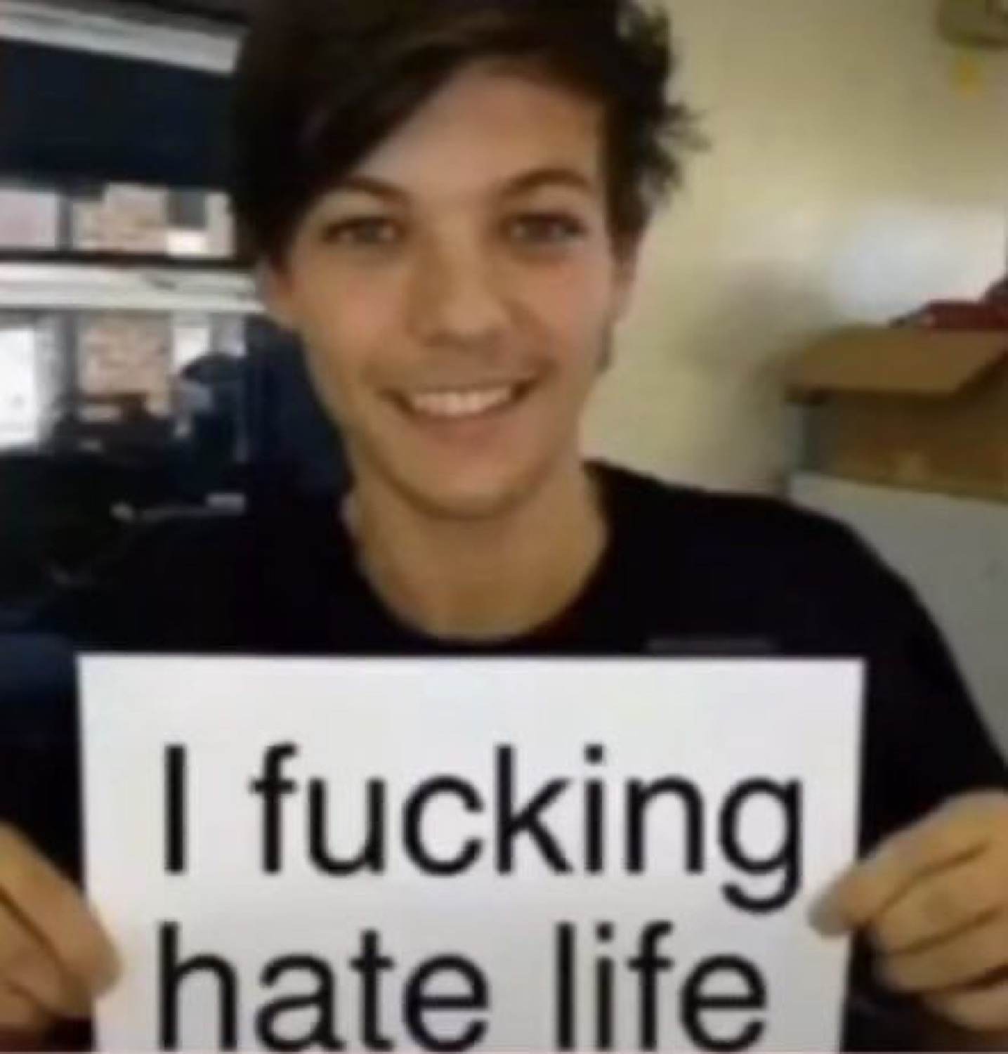 Louis Tomlinson grinning while holding a sign: 'I fucking hate life'