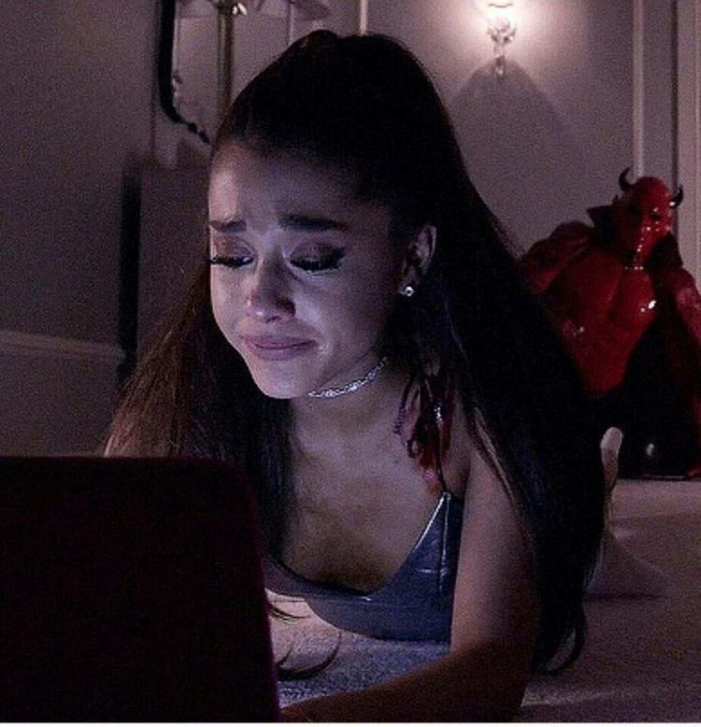 Young woman with a high ponytail crying at a laptop in a dim room as a red devil figure looms behind