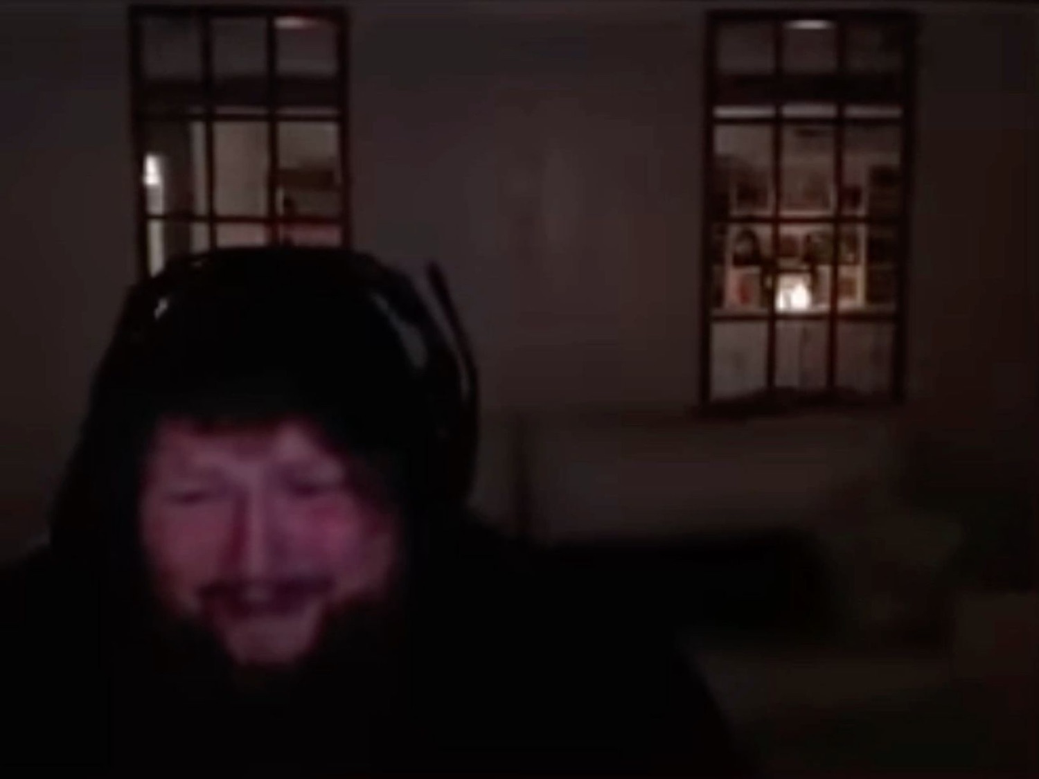 Man in a dark hoodie sitting in a dim room with a strained, uneasy grimace