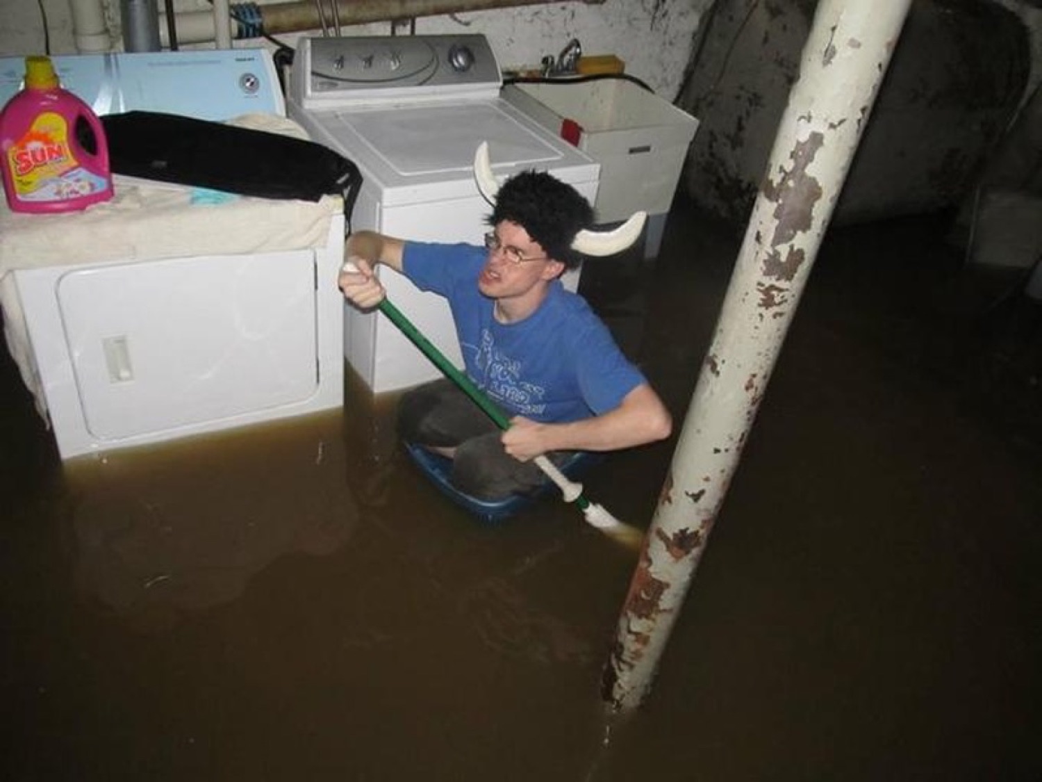 Guy in a viking helmet paddling a bin through a flooded basement past the washer and dryer