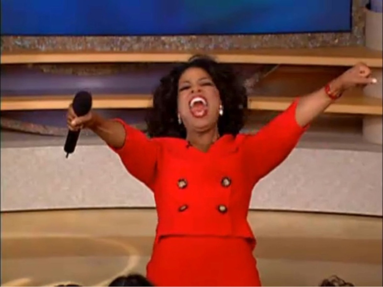 Talk show host in a red suit throwing her arms wide and screaming with pure excitement