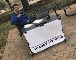 Man sitting outdoors at a table with a sign reading 'CHANGE MY MIND'