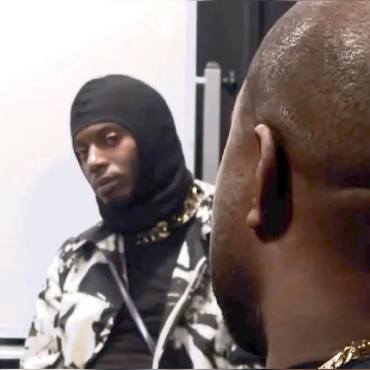 Man in a black balaclava giving a flat, deadpan stare at someone facing him