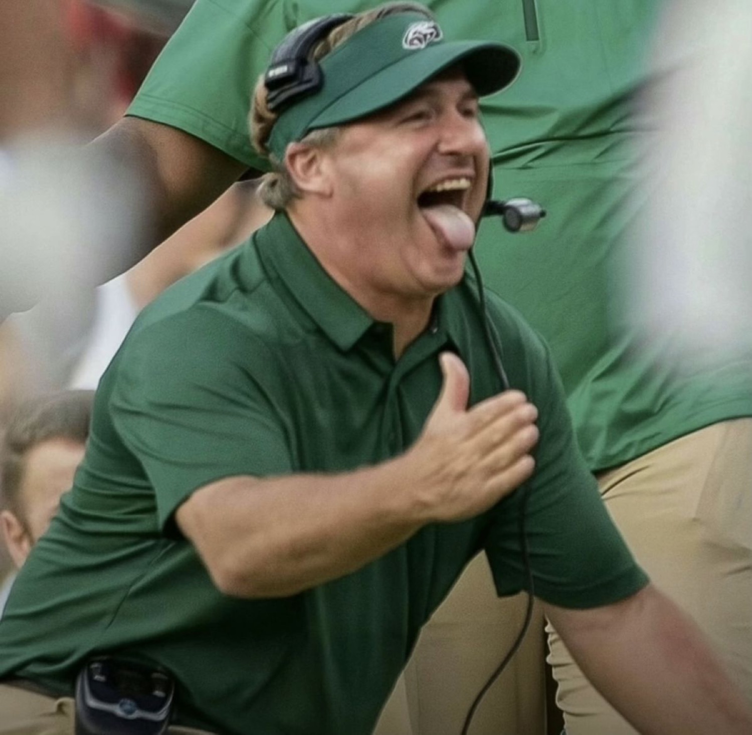 Football coach in a green polo screaming with his tongue out and fist to his chest in intense sideline hype