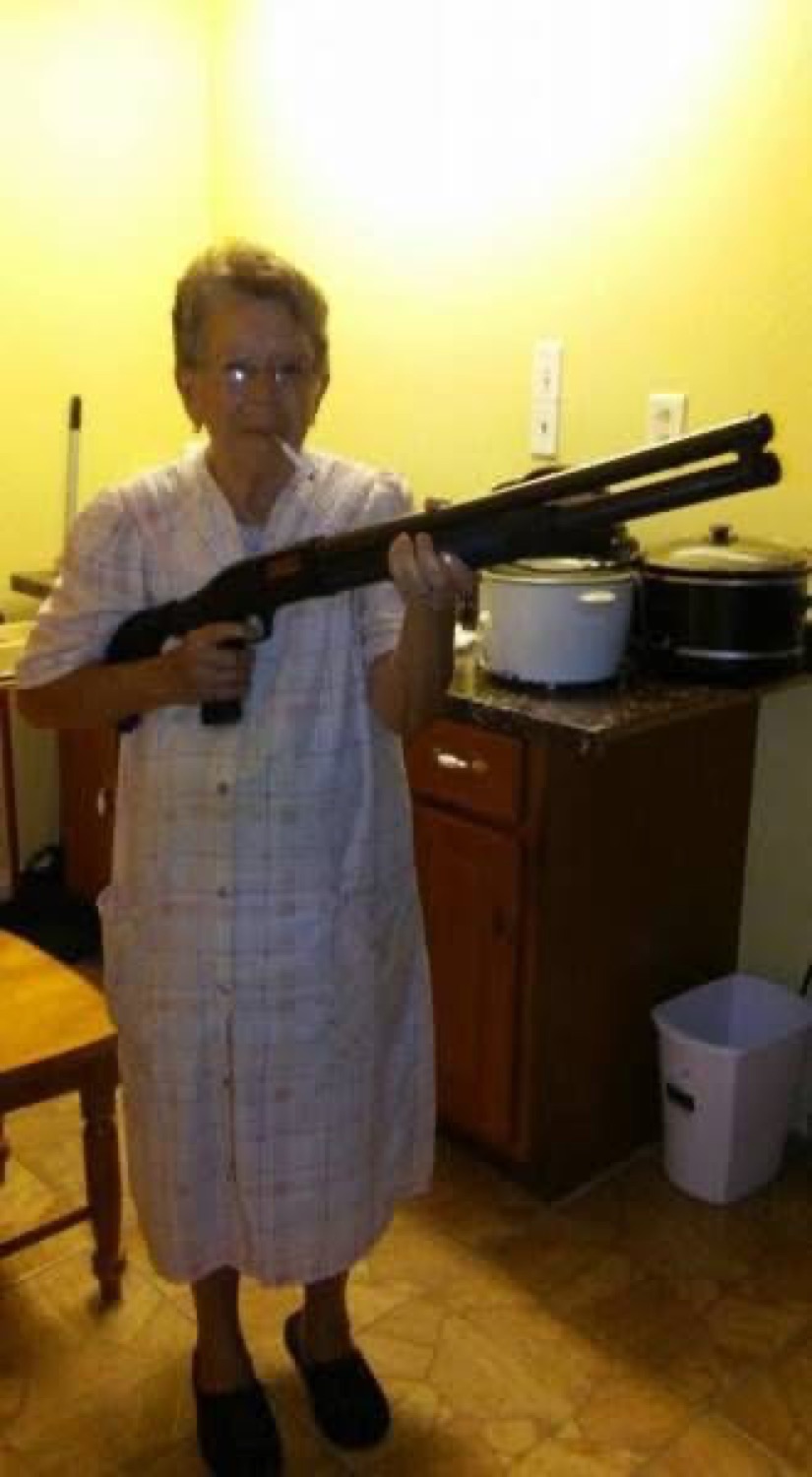 Elderly woman in a housedress standing in her kitchen holding a shotgun with total calm