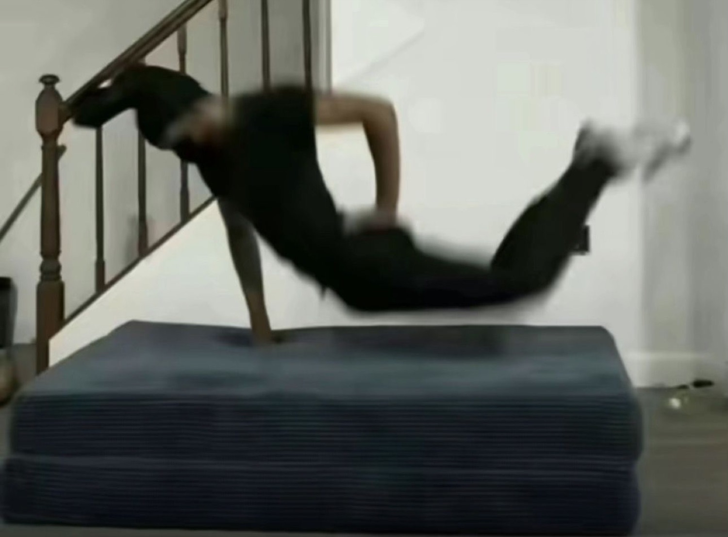 Person in all black flailing mid-air off a mattress at the bottom of a staircase