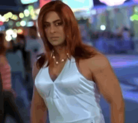 Muscular man in a long red wig and white top strutting down a street