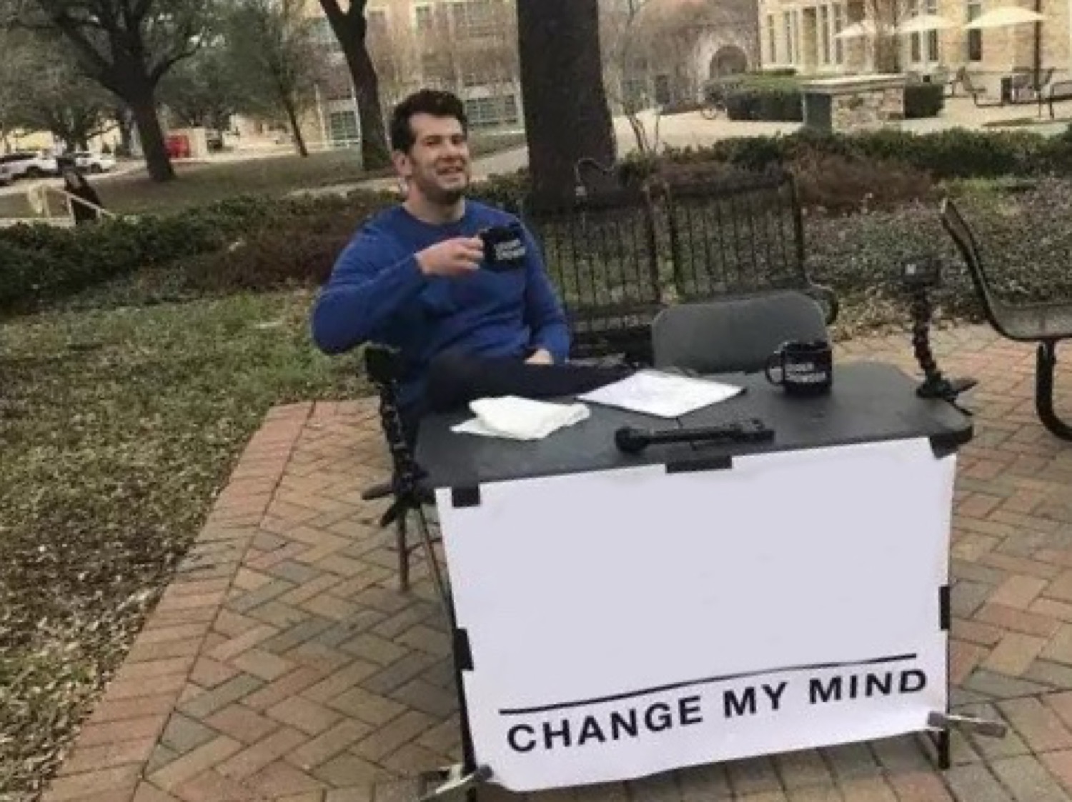 Man sitting at an outdoor table with a 'CHANGE MY MIND' sign, smug and ready to debate