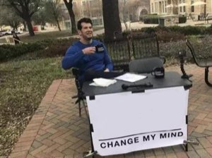 Man sitting at an outdoor table with a 'CHANGE MY MIND' sign, smug and ready to debate
