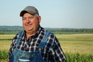 Farmer in overalls and a cap standing in a cornfield with a content, knowing smile