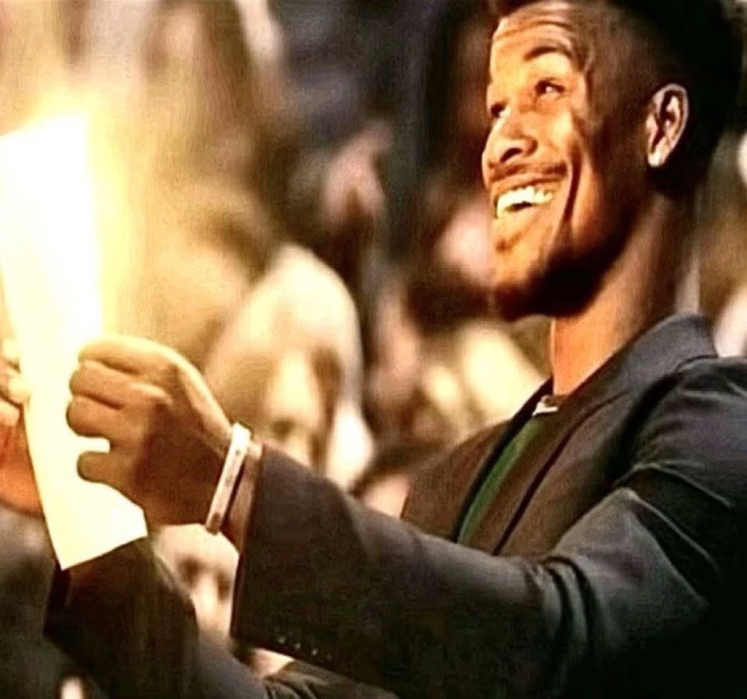 Man in a suit grinning widely while holding a piece of paper in a crowd