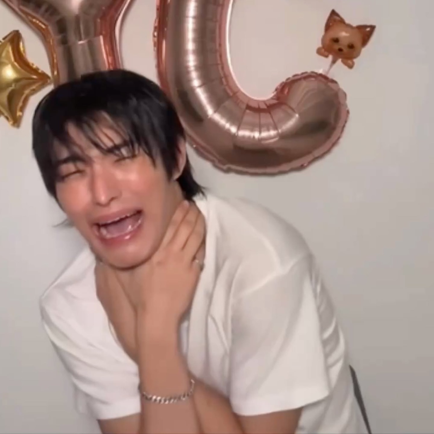 Guy in a white tee ugly-crying under birthday balloons, hand on his neck