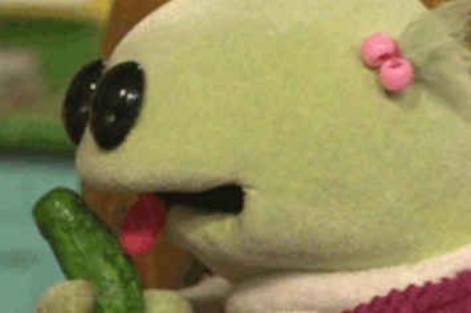 Green puppet with beady eyes and tongue out chewing on a pickle