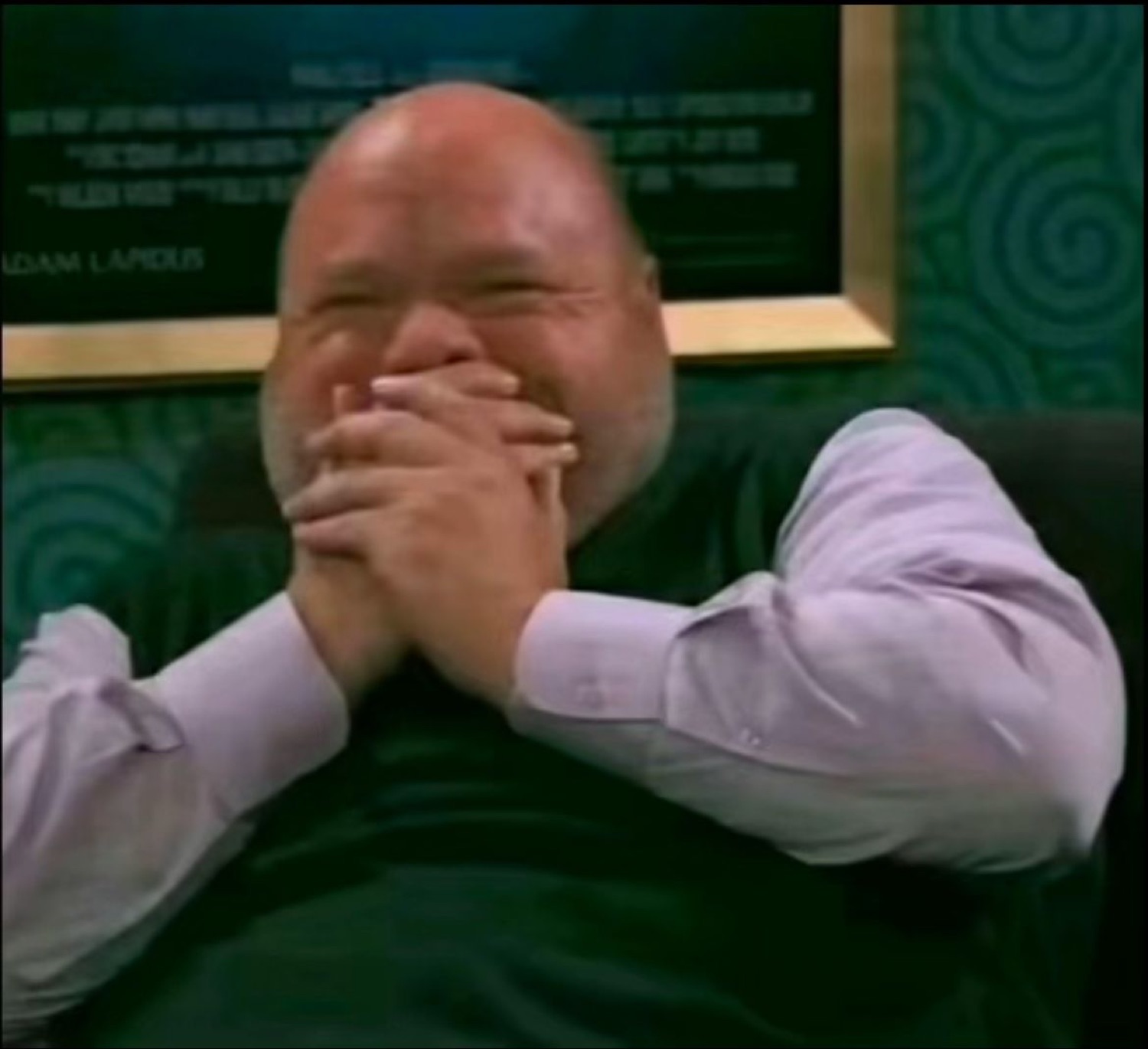 Bald man on a talk show barely holding it together, covering his face with both hands while laughing