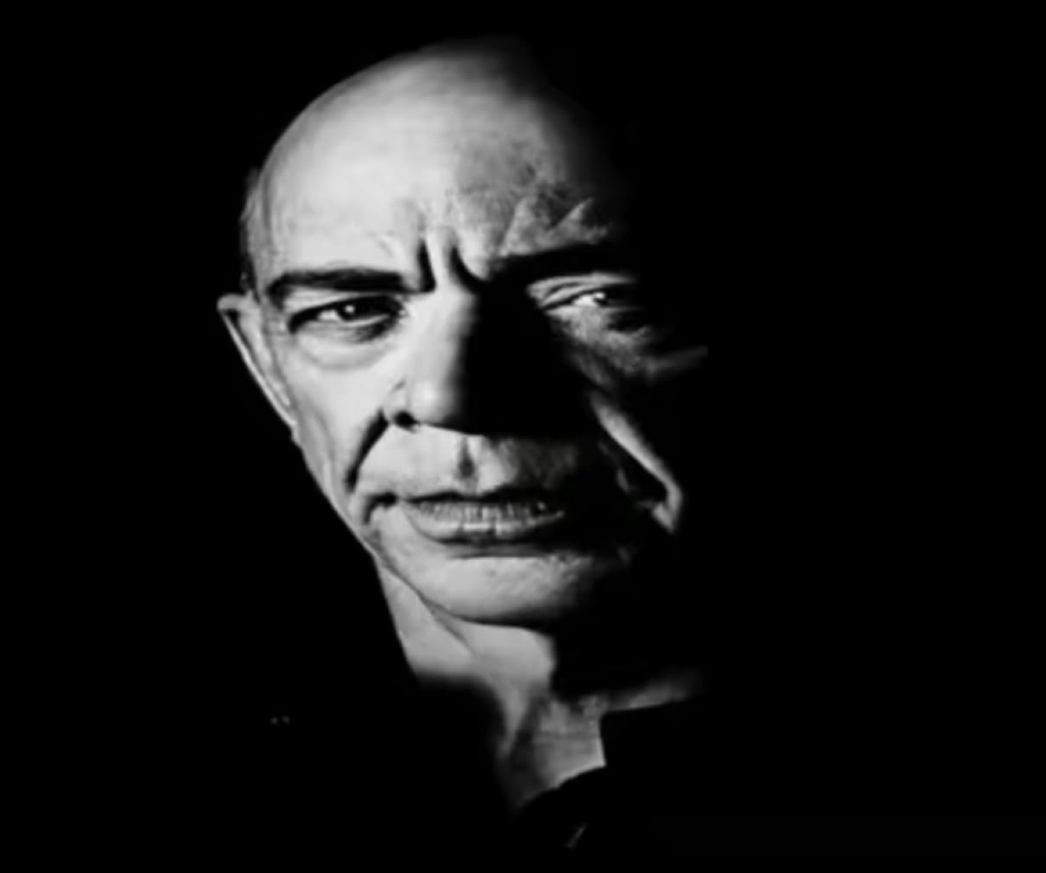 Bald man lit dramatically against black, scowling with a menacing, unimpressed glare