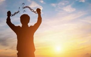 Silhouette of a person breaking chains with arms raised against a bright sunrise