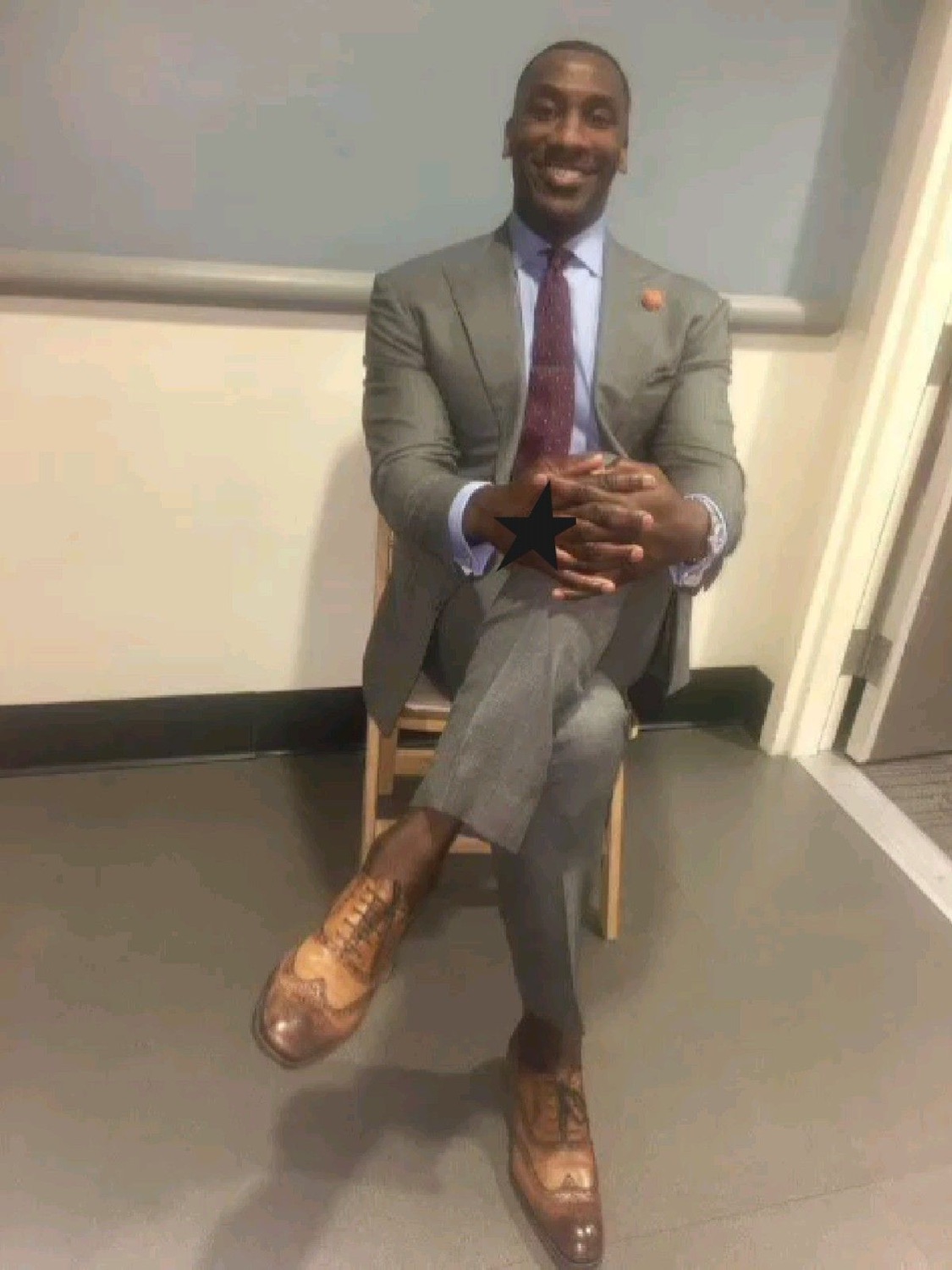 Man in a grey suit lounging cross-legged in a chair with a confident, pleased grin