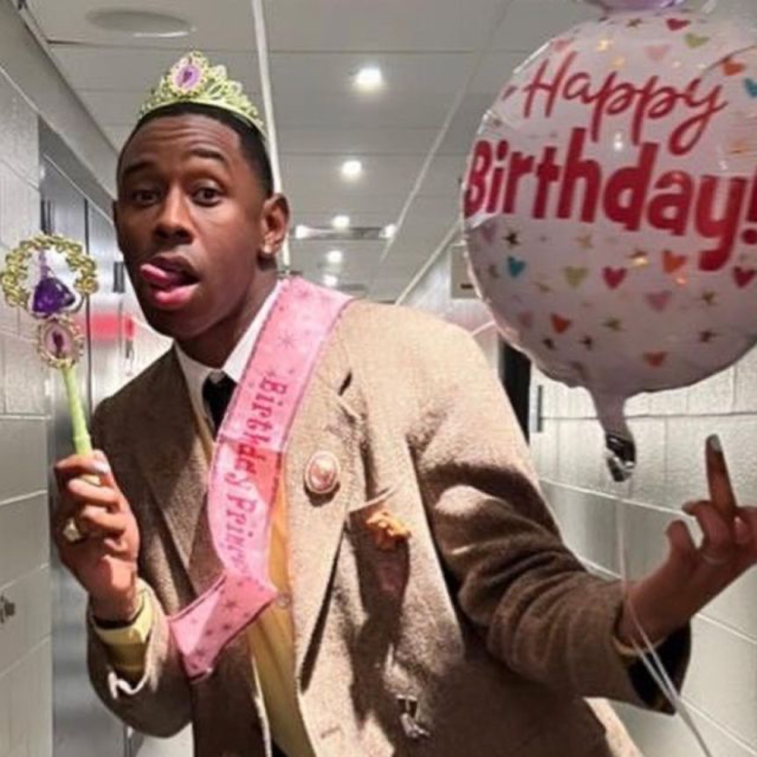 Tyler, the Creator in a tiara and birthday sash with a balloon, sticking his tongue out