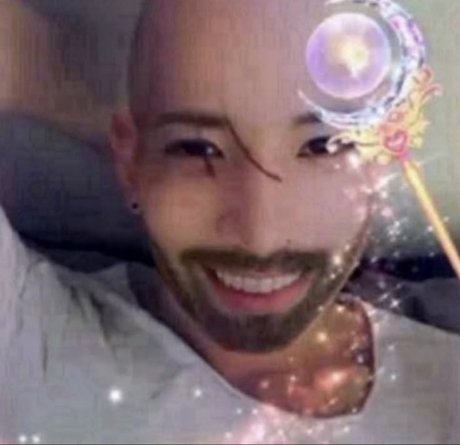 Bald bearded guy grinning in bed under a glittery magic-wand sparkle filter