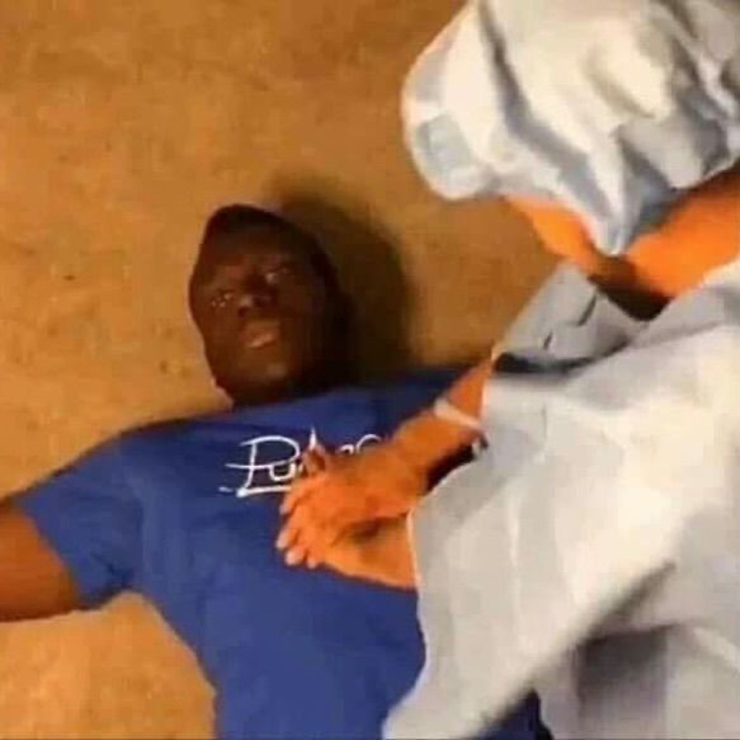 Guy in a blue tee lying flat on the floor while gloved hands tuck a sheet over him