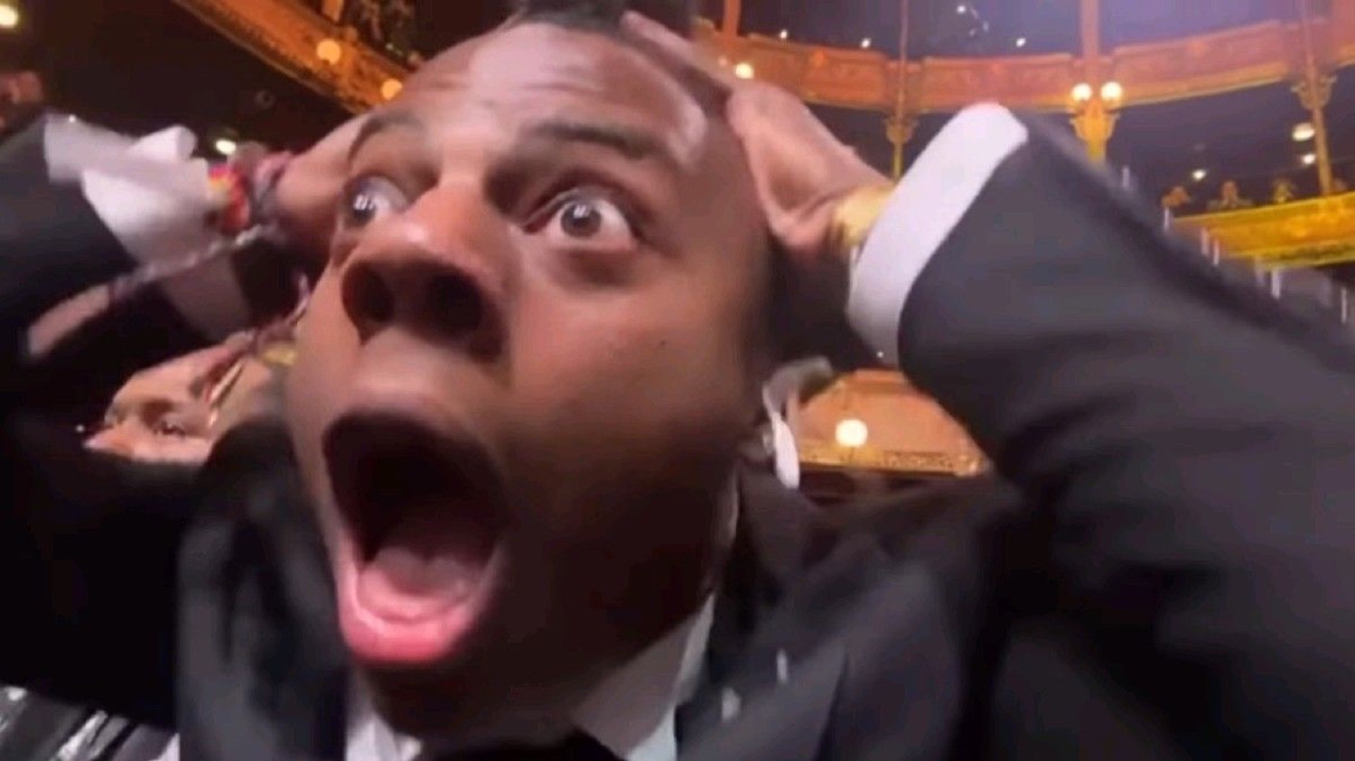 Man in a tuxedo gripping his head and screaming in wide-eyed shock at an event