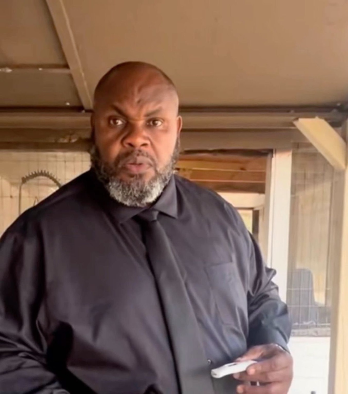 Bald bearded man in a black shirt scowling intensely with a phone in his hand