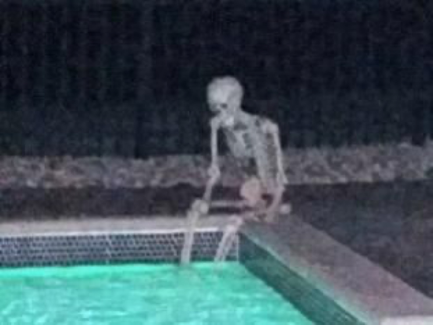 Skeleton sitting alone at the edge of a pool at night