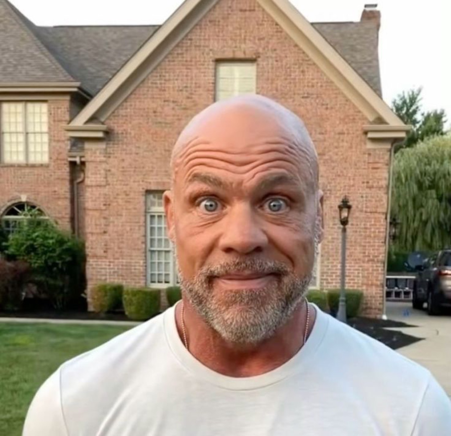 Bald muscular man in a white tee making a wide-eyed, startled awkward face outside a house