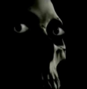 Dark moody face emerging from shadows with wide hollow eyes and a grimacing open mouth