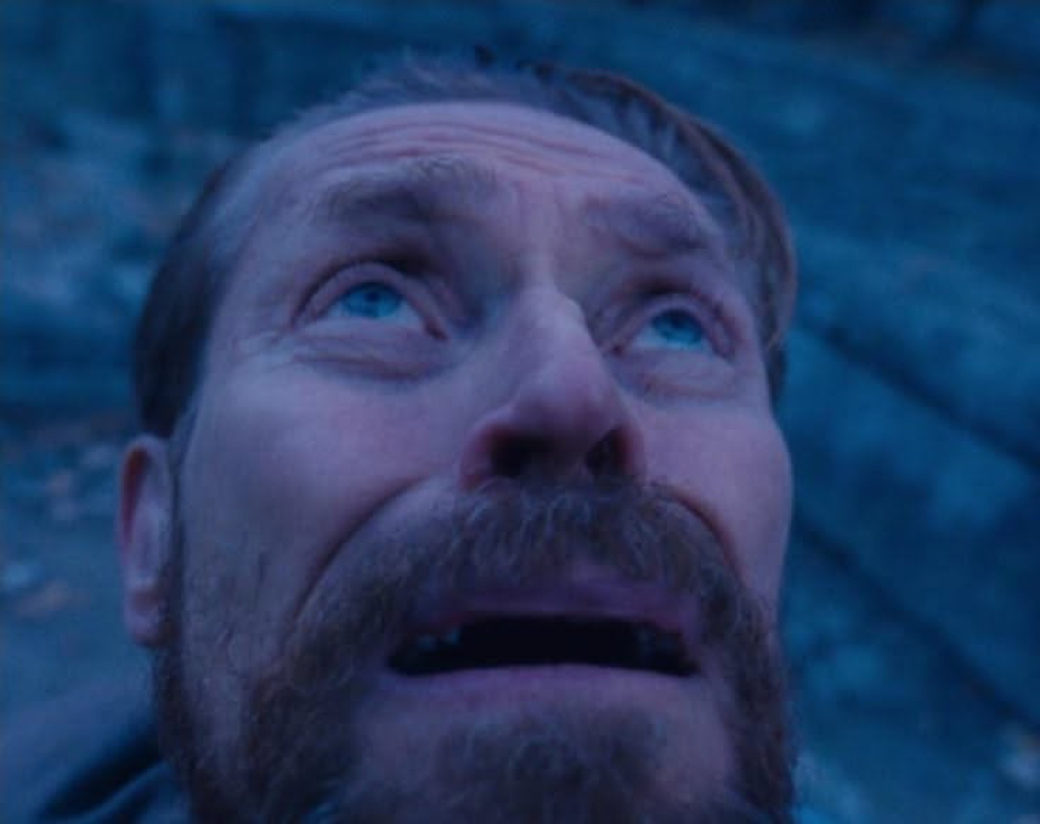 Bearded man staring upward in the cold blue light with terrified, teary eyes