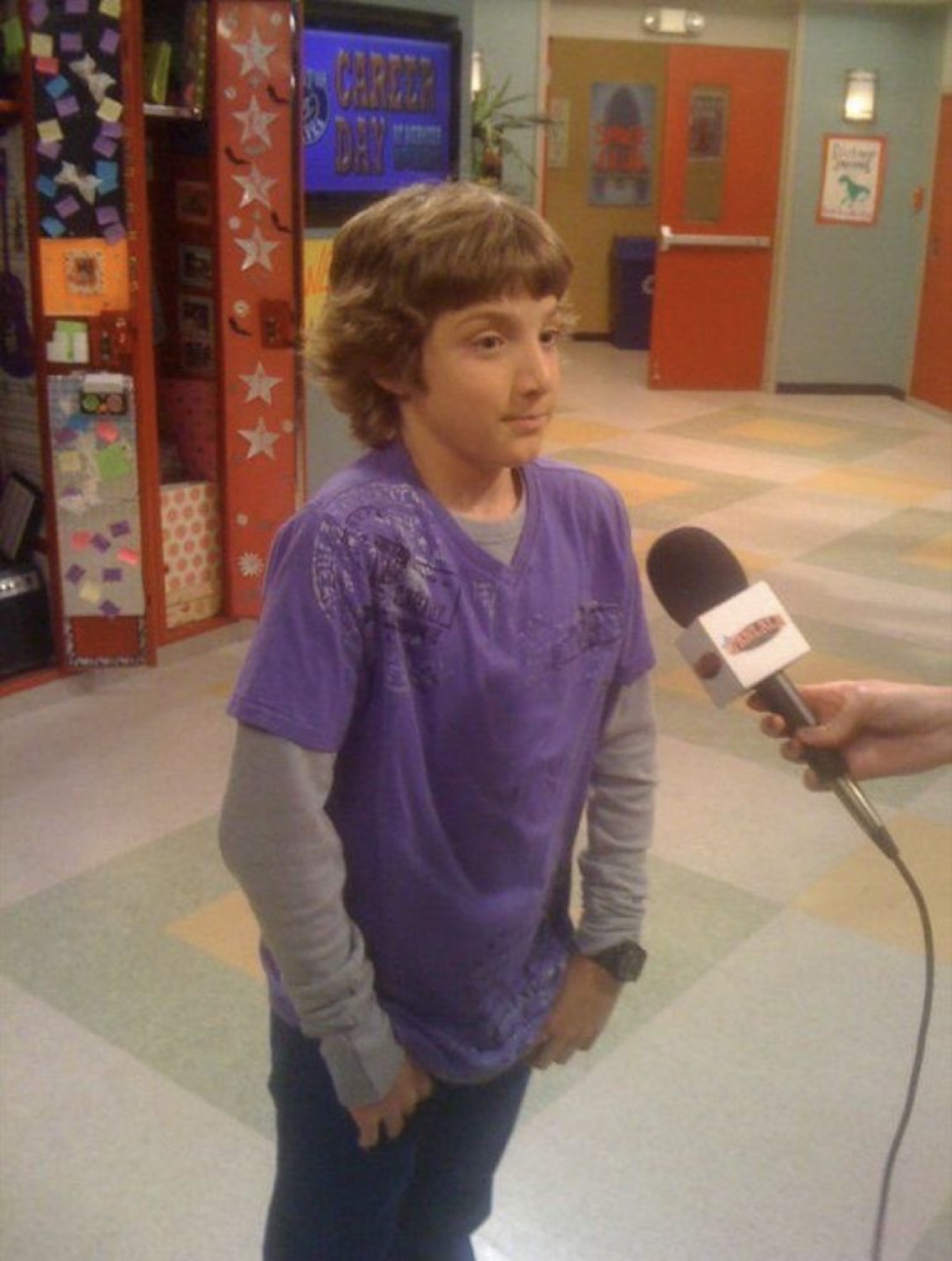 Teen boy in a purple shirt being interviewed with a mic in a school hallway, looking mildly blank