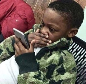 Little kid in camo staring at a phone with a hand clapped over his mouth in shock