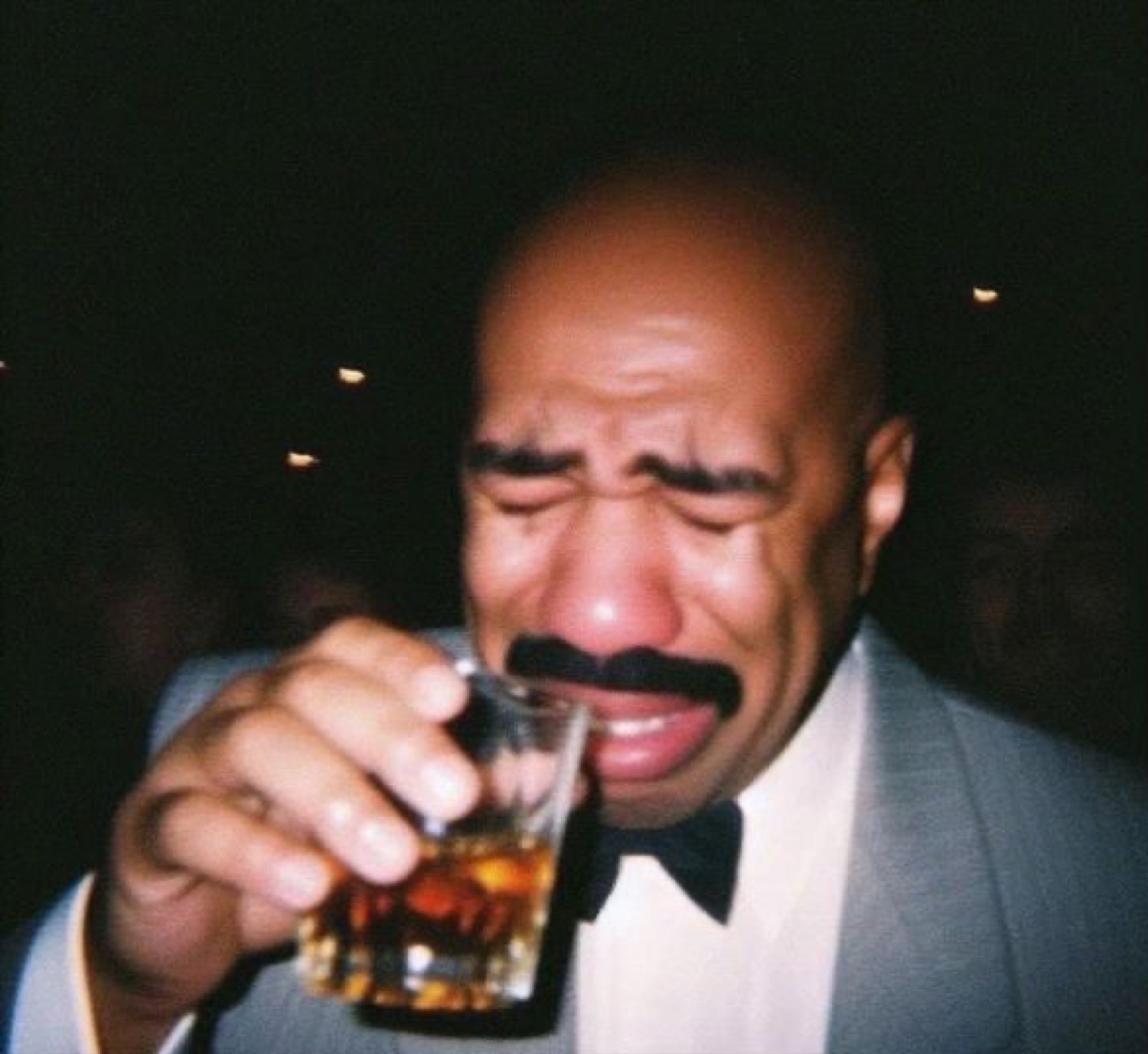 Steve Harvey in a tux, face scrunched up crying dramatically over a drink