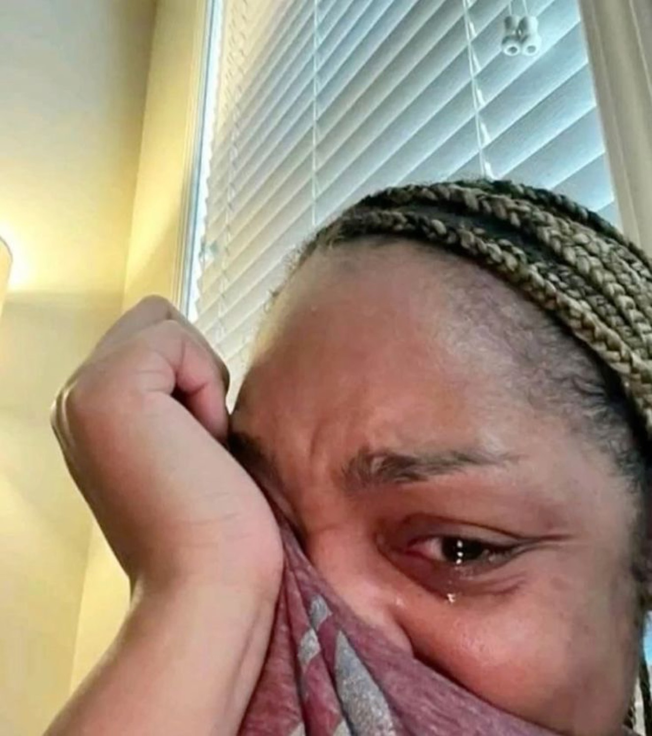 Woman wiping a tearful eye against a window blind, quietly emotional