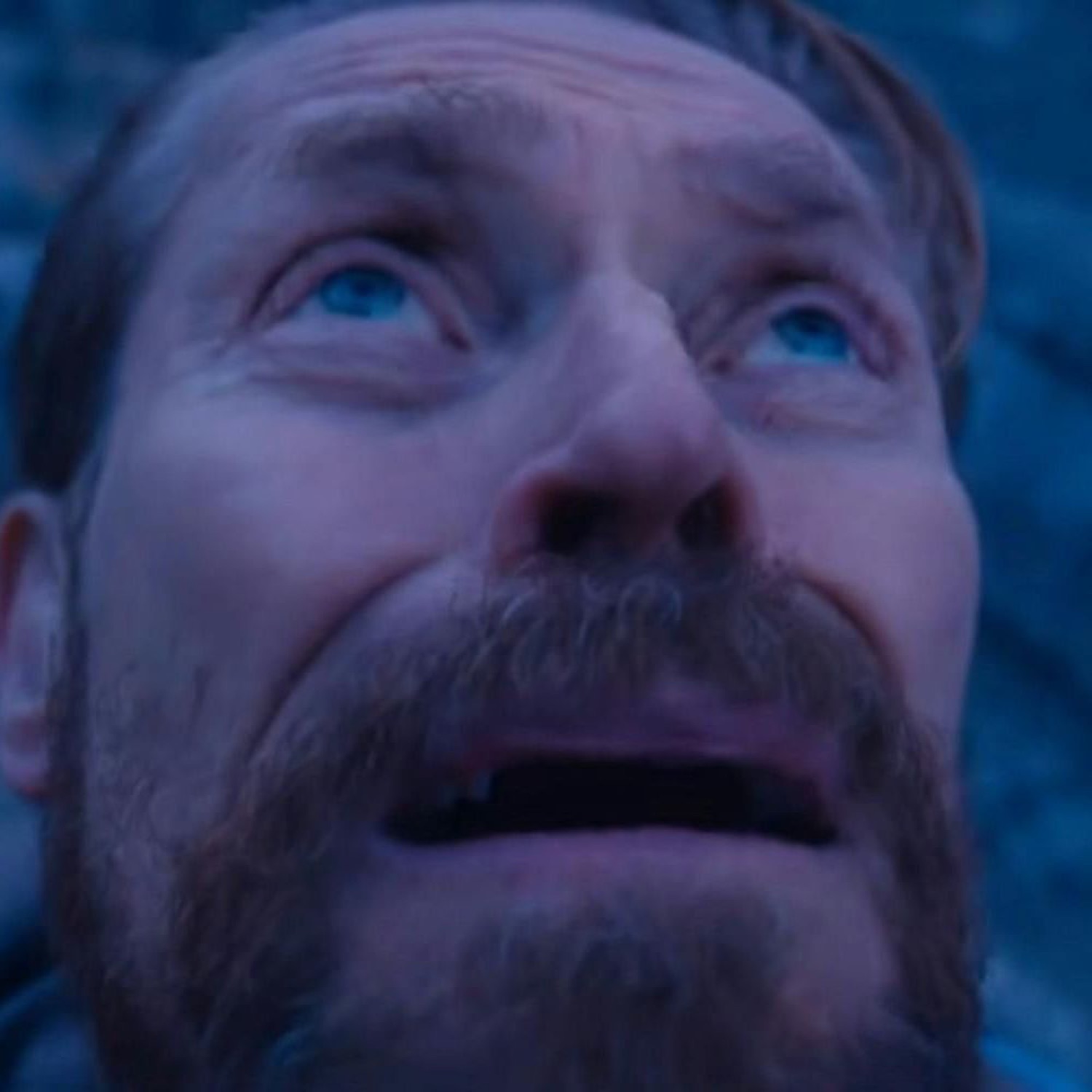 Bearded man gazing upward with wide, terrified, teary eyes in dim blue light