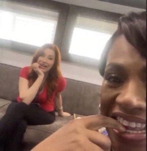 Two women laughing and smiling together on a couch, one leaning in close to the camera