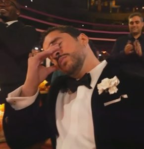 Man in a tuxedo at an awards show pressing his hand to his face in disbelief