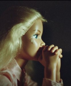 Doll with a human-like face resting on clasped hands in a pensive, prayerful pose