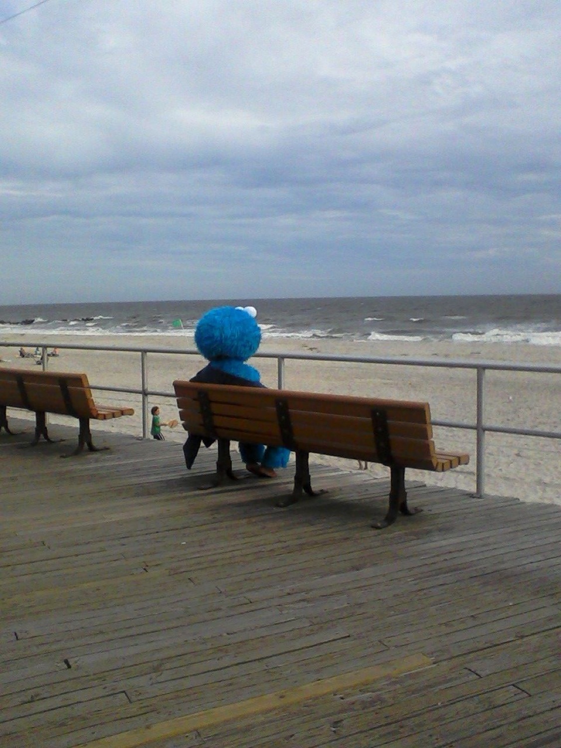 Cookie Monster costume sitting alone on a boardwalk bench staring out at the grey sea
