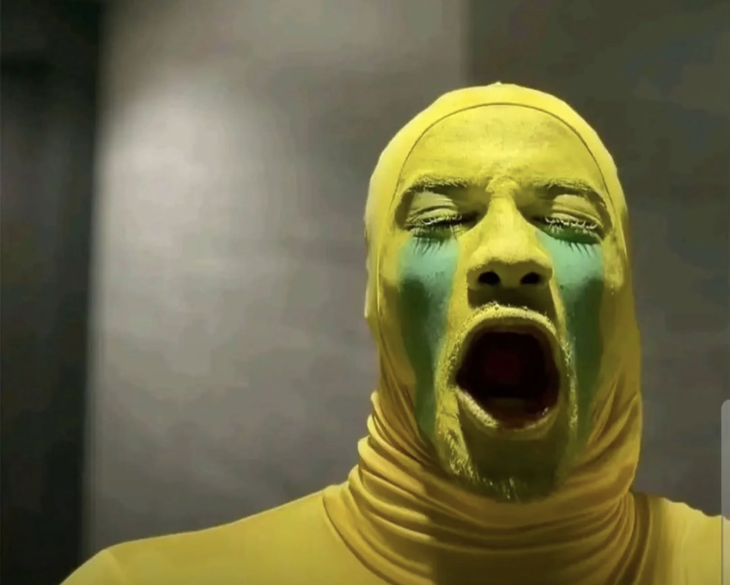 Person in a yellow full-body suit and yellow face paint screaming with eyes shut